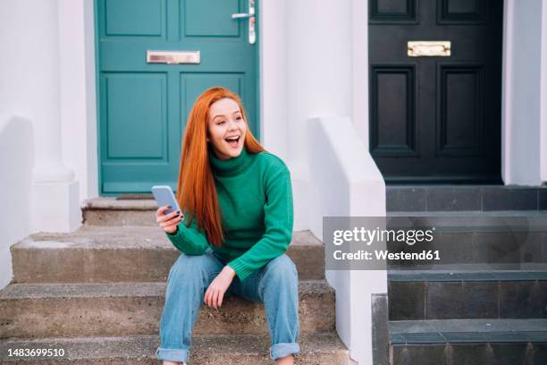 surprised redhead woman sitting with mobile phone outside house - house door stock pictures, royalty-free photos & images