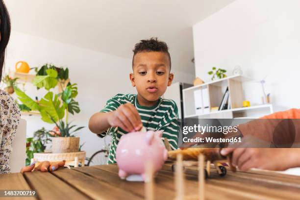 boy inserting coin into piggy bank at home - zakgeld stockfoto's en -beelden