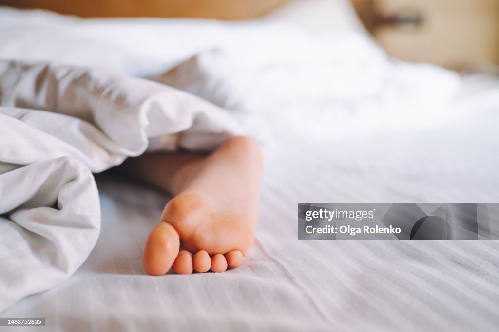 Innocent kid dream, pure child foot protruded from the blanket. Close up cute child barefoot stick out of white linen on bed. Copy space