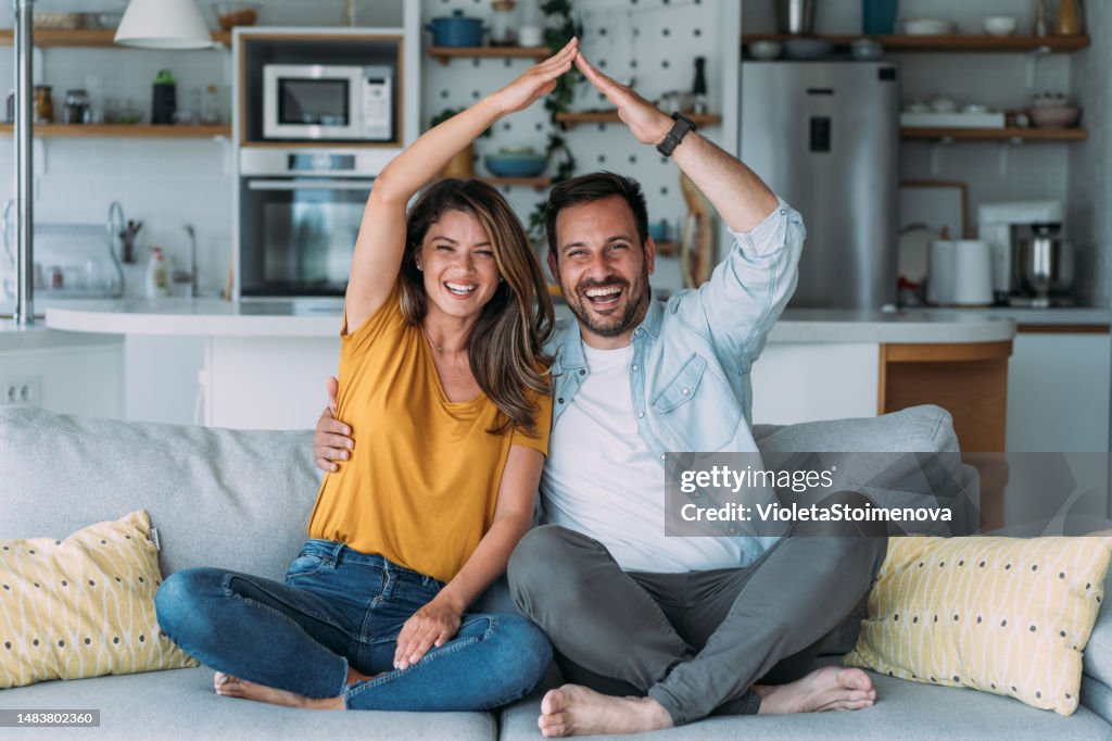 Happy couple making roof figure with their arms in the living room.