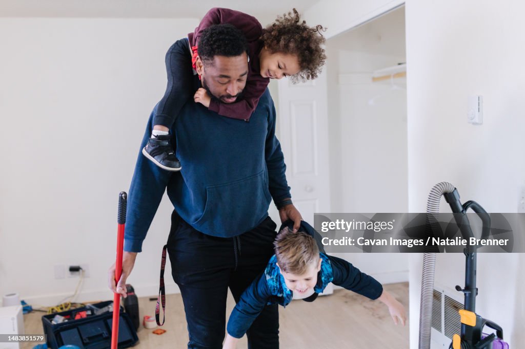 Black dad holding two kids while cleaning the house