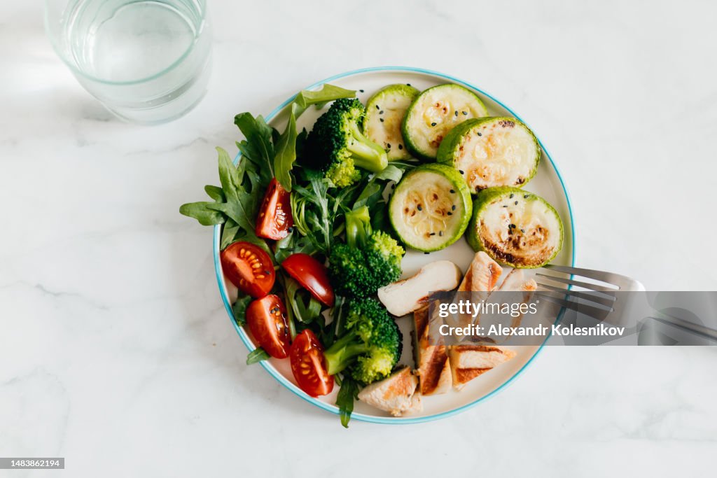 Grilled chicken meat and fresh vegetables tomato, broccoli, zucchini and arugula on plate, glass with water on white background. Organic balanced food. Top view
