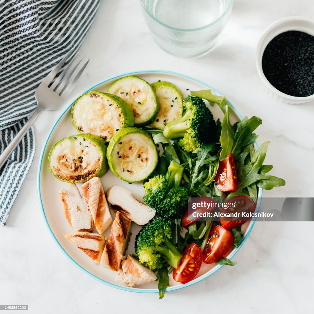 Grilled chicken meat and fresh vegetables tomato, broccoli, zucchini and arugula on plate, glass with water on white background. Organic balanced food. Top view