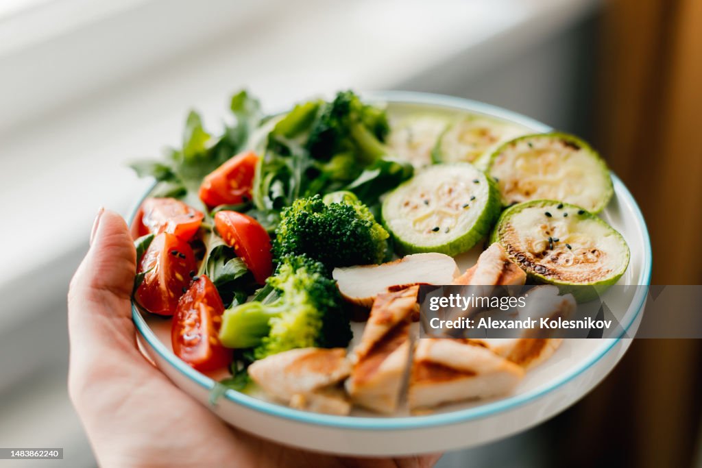 Grilled chicken meat and fresh vegetable tomato, broccoli, zucchini and arugula on plate in woman's hand. Organic balanced food