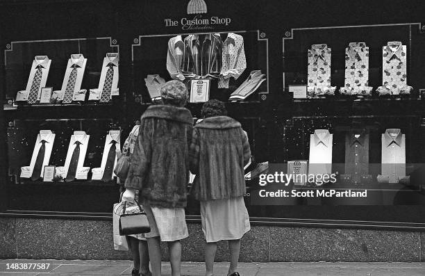View of two women, both in fur coats, as they stand in front of a window display of dress shirts outside of Sak's Fifth Avenue department store on...