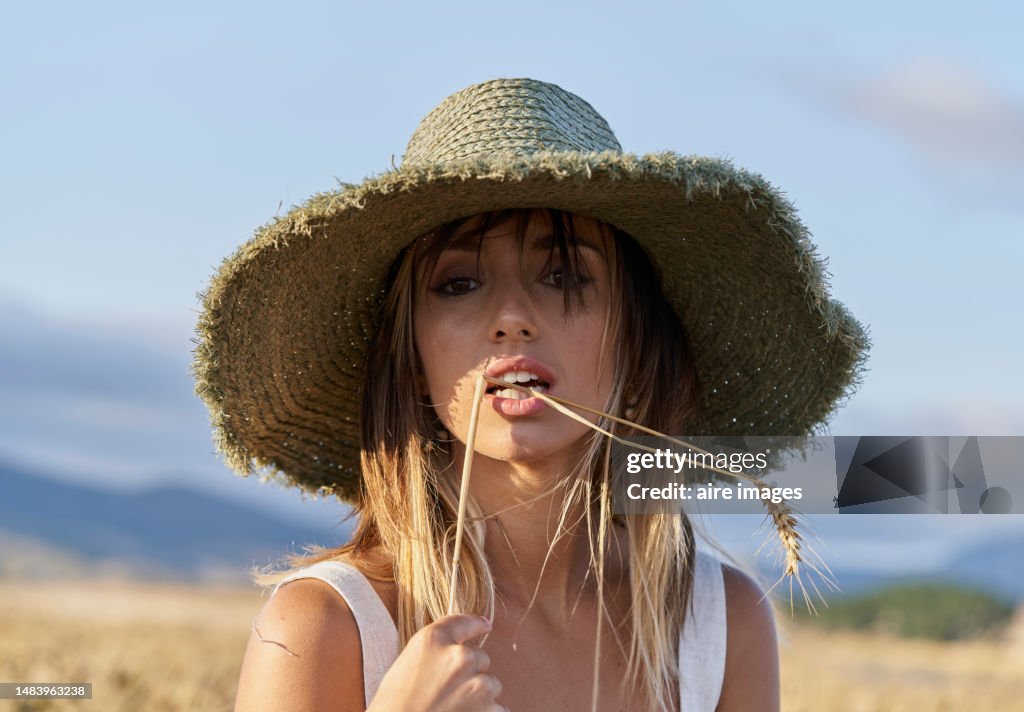 Frontal close-up view of a beautiful woman standing in a field looking sensually at the camera holding and biting a wheat.
