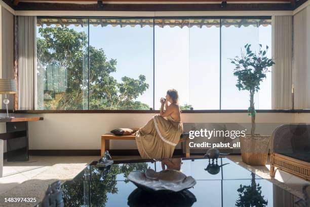 mature woman drinking by hotel window - fashion for peace stockfoto's en -beelden