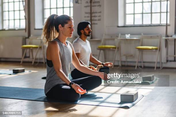 an adult  caucasian brunette female meditating in a yoga class next to an indian male in a beautiful yoga studio - yoga studio stock pictures, royalty-free photos & images