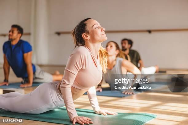 uma fêmea atraente com olhos fechados fazendo uma pose de cobra durante uma aula de ioga em um belo estúdio com luz solar natural - yoga studio - fotografias e filmes do acervo