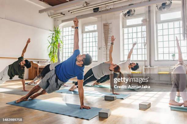 diverse yoga class participants doing a side plank on their yoga mats in a beautiful yoga studio with big windows - pilates stock pictures, royalty-free photos & images