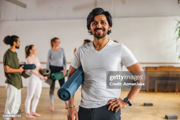 adult indian  male yoga instructor smiling at the camera and holding a yoga mat under his arm while his clients are having a conversation in the background - professor de ioga imagens e fotografias de stock