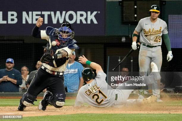 Conner Capel of the Oakland Athletics slides into home plate as Jonah Heim of the Texas Rangers tries to catch the ball at Globe Life Field on April...