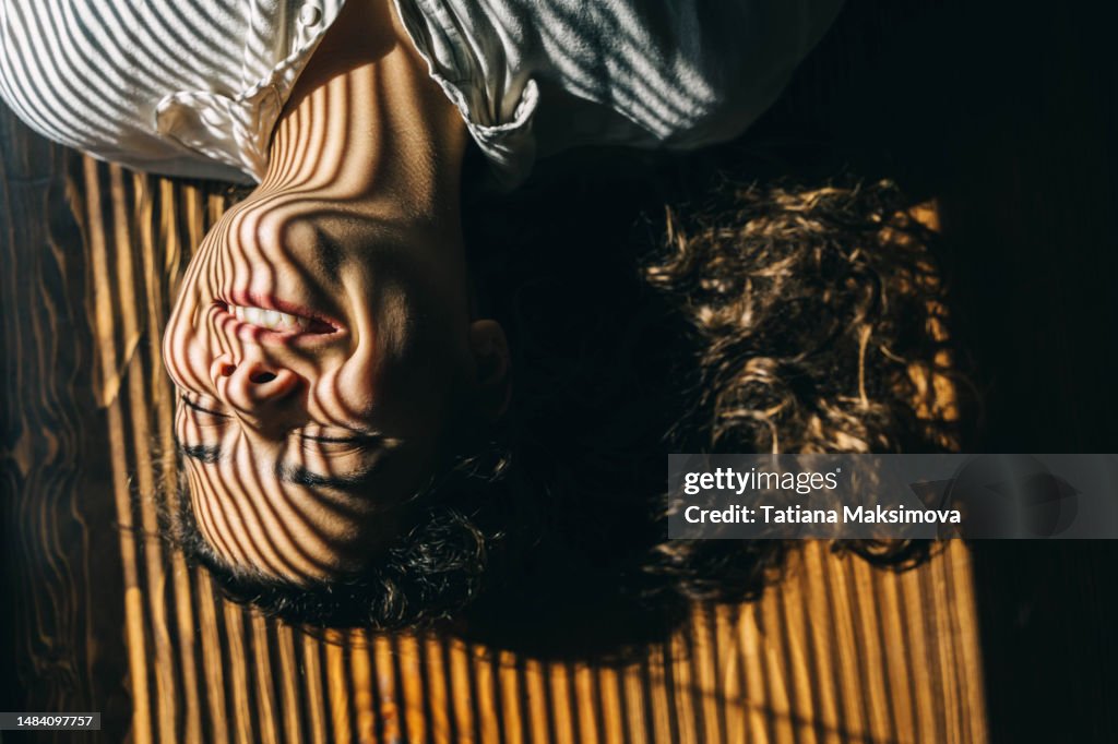 Beautiful young woman enjoys the sunlight lying on the floor. Striped light from blinds. Top view.