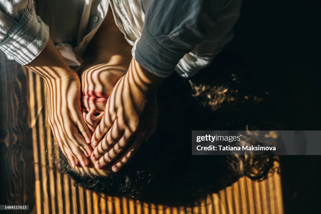 A depressed woman lies on wooden floor and closes her face with hand, striped from blinds on face. Top view.