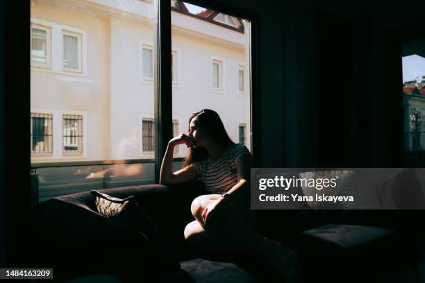 a tired and depressed mediterranean woman is sitting on the sofa near the window at home - toxisch sociaal concept stockfoto's en -beelden