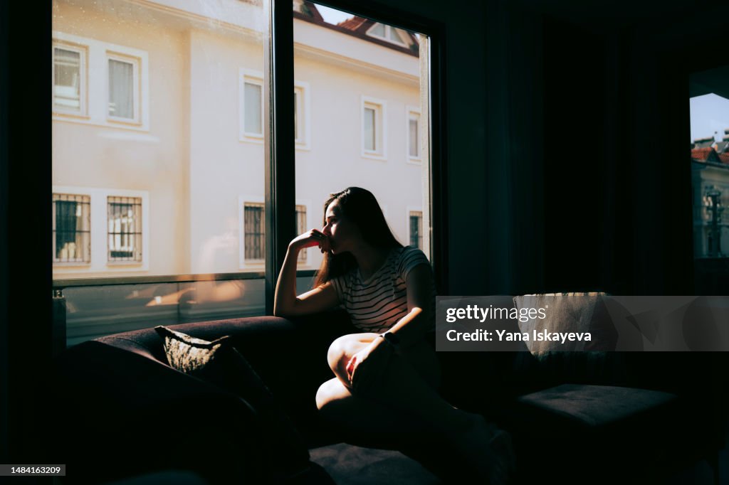 A tired and depressed Mediterranean woman is sitting on the sofa near the window at home
