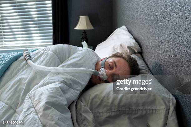 a 28-year-old man sleeps with a cpap machine in bed. the machine helps him breathe better by delivering air pressure through a mask, improving sleep quality and reducing snoring - 55 year old male stockfoto's en -beelden