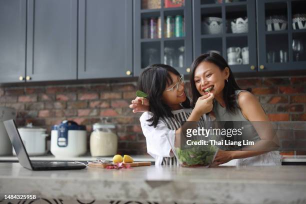a cheerful asian mother and daughter are using a laptop computer in the kitchen and having fun while mixing salad in a bowl. - asian mother cooking imagens e fotografias de stock