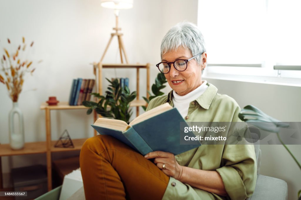 Senior woman sitting on sofa in the living room, relaxing and reading book at home