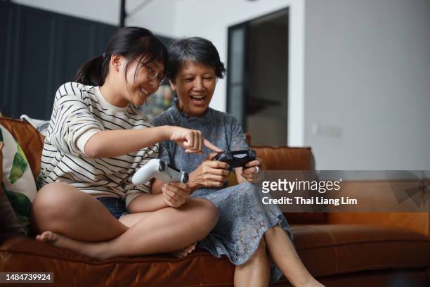 un'allegra nonna asiatica e sua nipote sono sedute sul divano in salotto, imparando e divertendosi a giocare ai videogiochi insieme a casa. questa scena commovente incarna il concetto di pensionamento felice. - scene di videogiochi foto e immagini stock