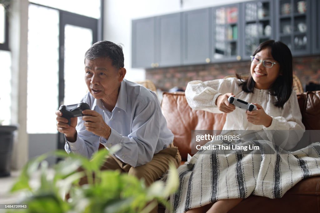 Un alegre abuelo asiático y su nieta están sentados en el sofá de la sala de estar, divirtiéndose jugando videojuegos juntos en casa. Esta conmovedora escena encarna el concepto de jubilación feliz