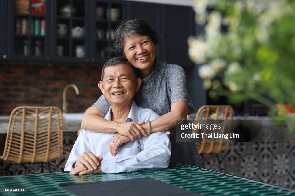 Pareja de ancianos asiáticos abrazados frente a su hogar, encarnando el concepto de estilo de vida saludable para personas mayores