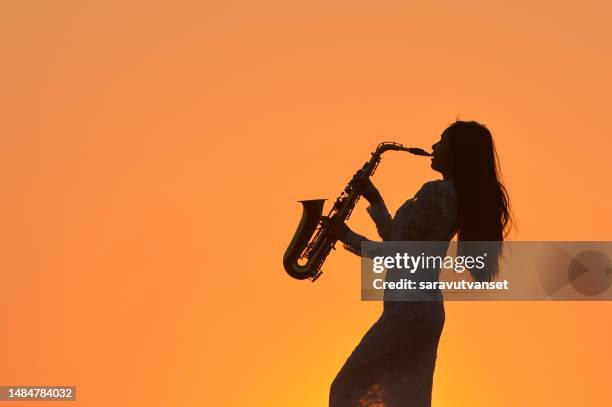 silhouette of a female saxophonist playing the saxophone by a lake at sunset, thailand - sassofono foto e immagini stock