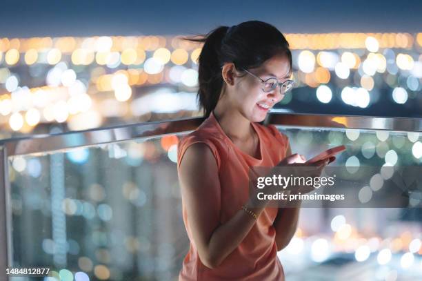 gen z young asian woman looking at smartphone surfing on the net while relaxing on the balcony in the evening. illuminated cityscape during twilight in background. lifestyle and technology - financial advisor virtual stock pictures, royalty-free photos & images