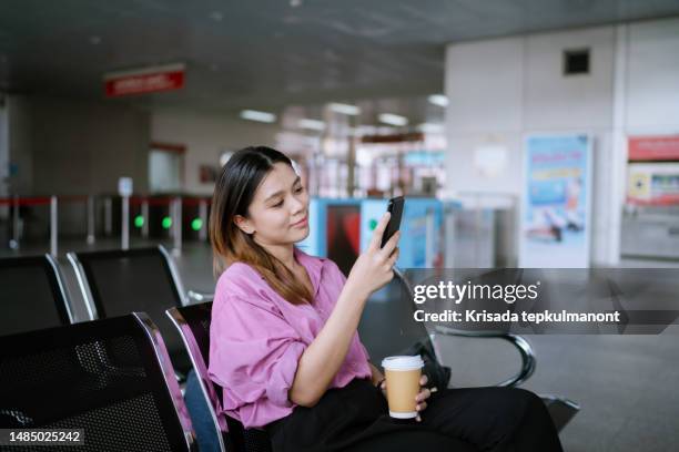 asian woman using mobile phone and drinking coffee while waiting for commuter bus during business trip. - bus operator stock pictures, royalty-free photos & images