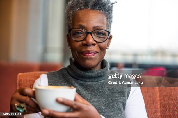 senior adult black female at a cafe smiling at the camera while holding a cup of delicious coffee in her hands - society of peace stock pictures, royalty-free photos & images