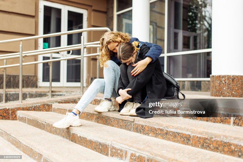 Unrecognizable female students sitting on the steps of the school, a friend supporting a frustrated disappointed and sad girl