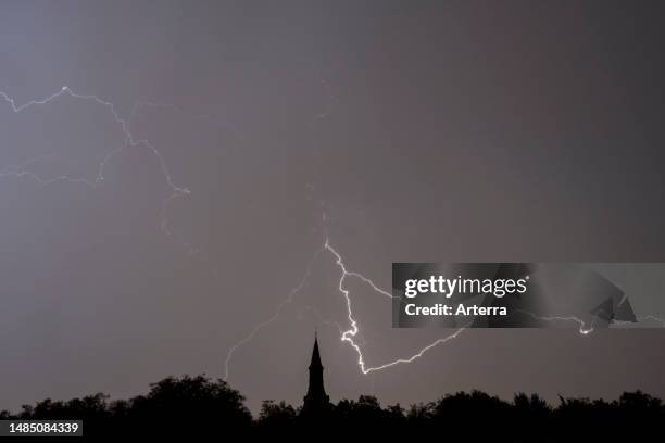 Rain falling and stroke of forked lightning during thunderstorm at night over church tower and trees.