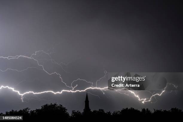 Rain falling and stroke of forked lightning during thunderstorm at night over church tower and trees.