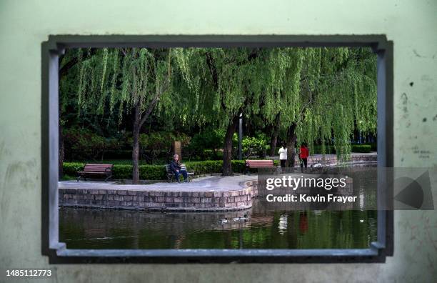 Man is seen through a window as he watches ducks pass by while sitting on a bench next to a lake at a local park in the evening on April 25, 2023 in...