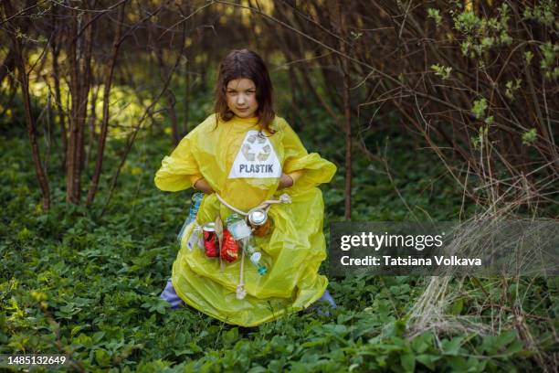 little kid in yellow raincoat posing and looking at camera while collecting garbage among trees - trash bag dress stock pictures, royalty-free photos & images