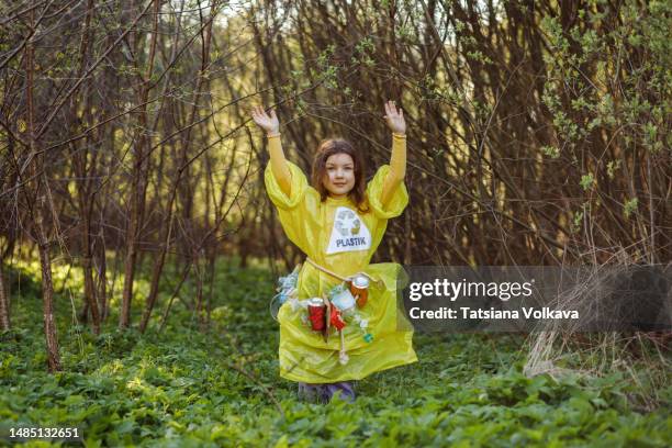 pre-adolescent girl wearing dress with recycling symbol standing among bushes with arms raised - trash bag dress stock pictures, royalty-free photos & images