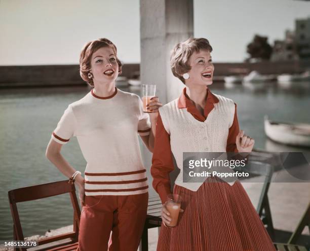 Vacation scene of two female fashion models at a quayside cafe wearing, on left, a white short sleeved sweater with red stripes and red trousers, on...