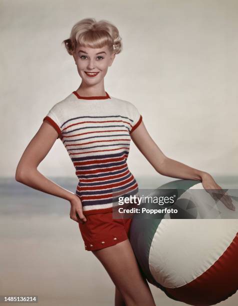 Posed studio portrait of a female fashion model wearing a short sleeved white knit top with red and blue horizontal stripes, she wears red shorts and...