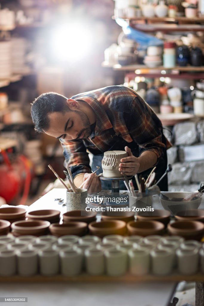 Young man potter making pattern on clay mug with special tool in pottery workshop, studio.