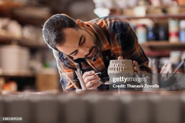 young man potter making pattern on clay mug with special tool in pottery workshop, studio. - moulant photos et images de collection