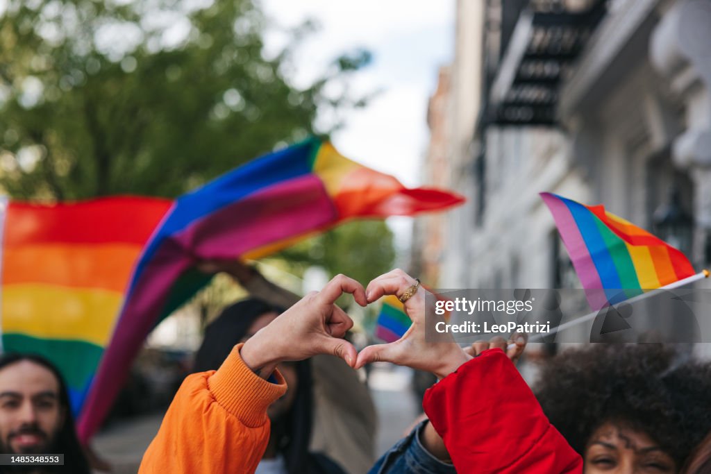 Genderqueer and non-binary friends making a heart shape against a rainbow flag