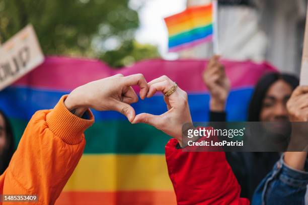 amici genderqueer e non binari che fanno una forma a cuore contro una bandiera arcobaleno - pride foto e immagini stock