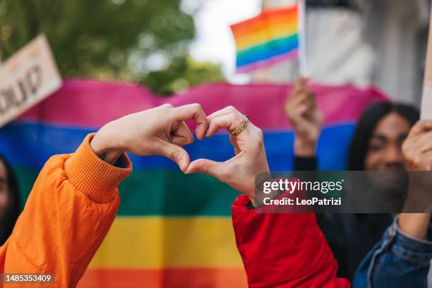 genderqueer and non-binary friends making a heart shape against a rainbow flag - lgbtqi stockfoto's en -beelden