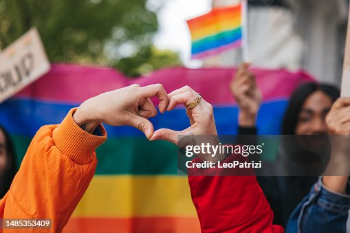 Genderqueer and non-binary friends making a heart shape against a rainbow flag Genderqueer and non-binary friends making a heart shape against a rainbow flag