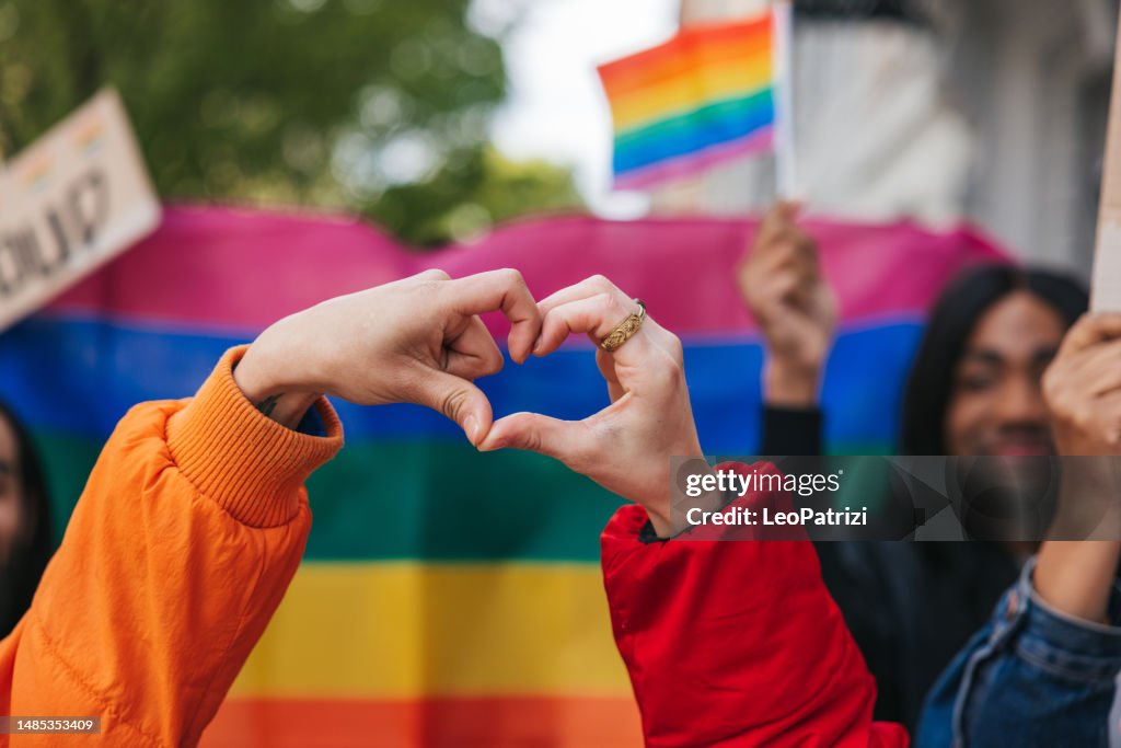 Genderqueer and non-binary friends making a heart shape against a rainbow flag