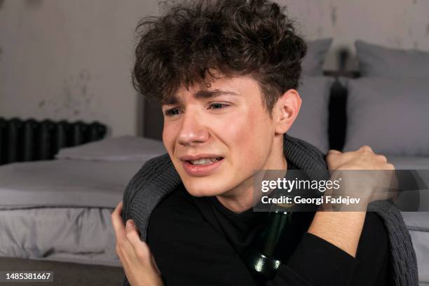 a severely depressed guy, sits on the floor covered with a blanket, with a bottle in hands. a young man in a hard mental condition. alcohol addiction. - fobie stockfoto's en -beelden