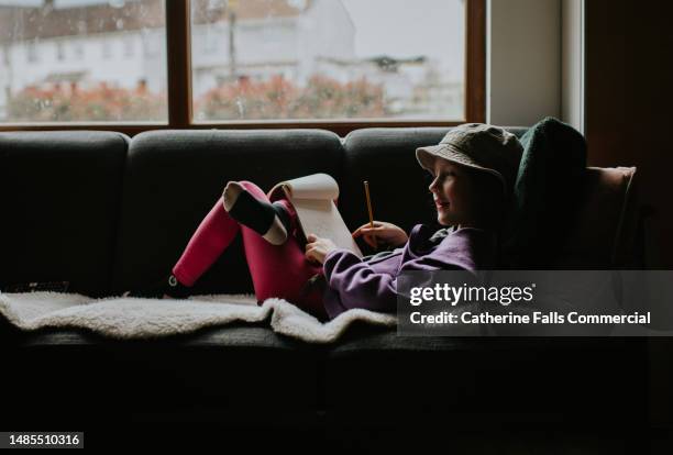 a young girl, wearing a hat, reclines on a sofa and doodles in a notebook with a pencil - liet stock pictures, royalty-free photos & images