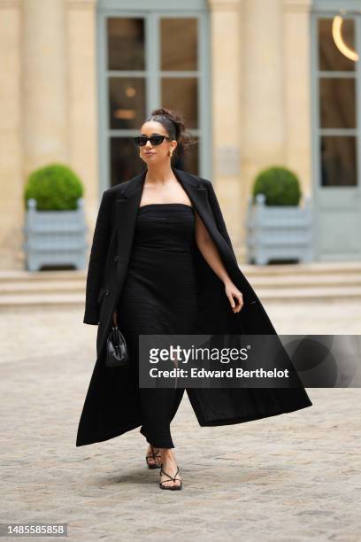 Lena Mahfouf wears sunglasses, a black long coat, an off-shoulder black maxi long slit dress, a black leather bag, black shoes, outside the COS show,...