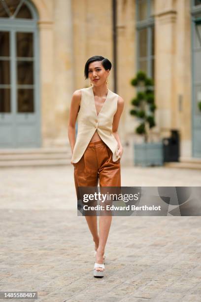 Molly Chiang wears golden earrings, a beige linen sleeveless jacket, brown leather embossed shorts, white platform shoes, outside the COS show, on...