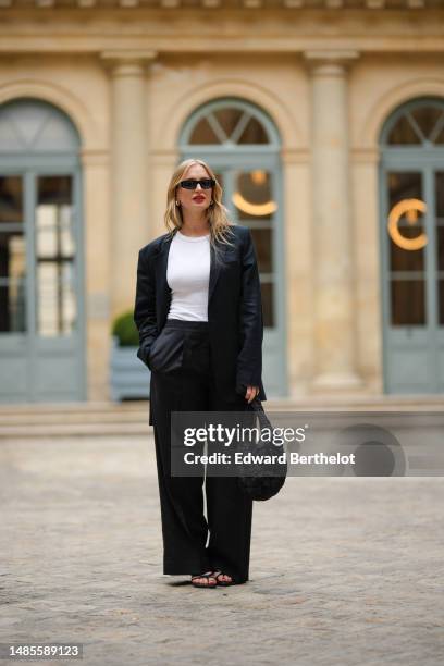 Guest wears sunglasses, earrings, a white t-shirt, a black oversize blazer jacket, black suit pants, a black puffy bag, outside the COS show, on...